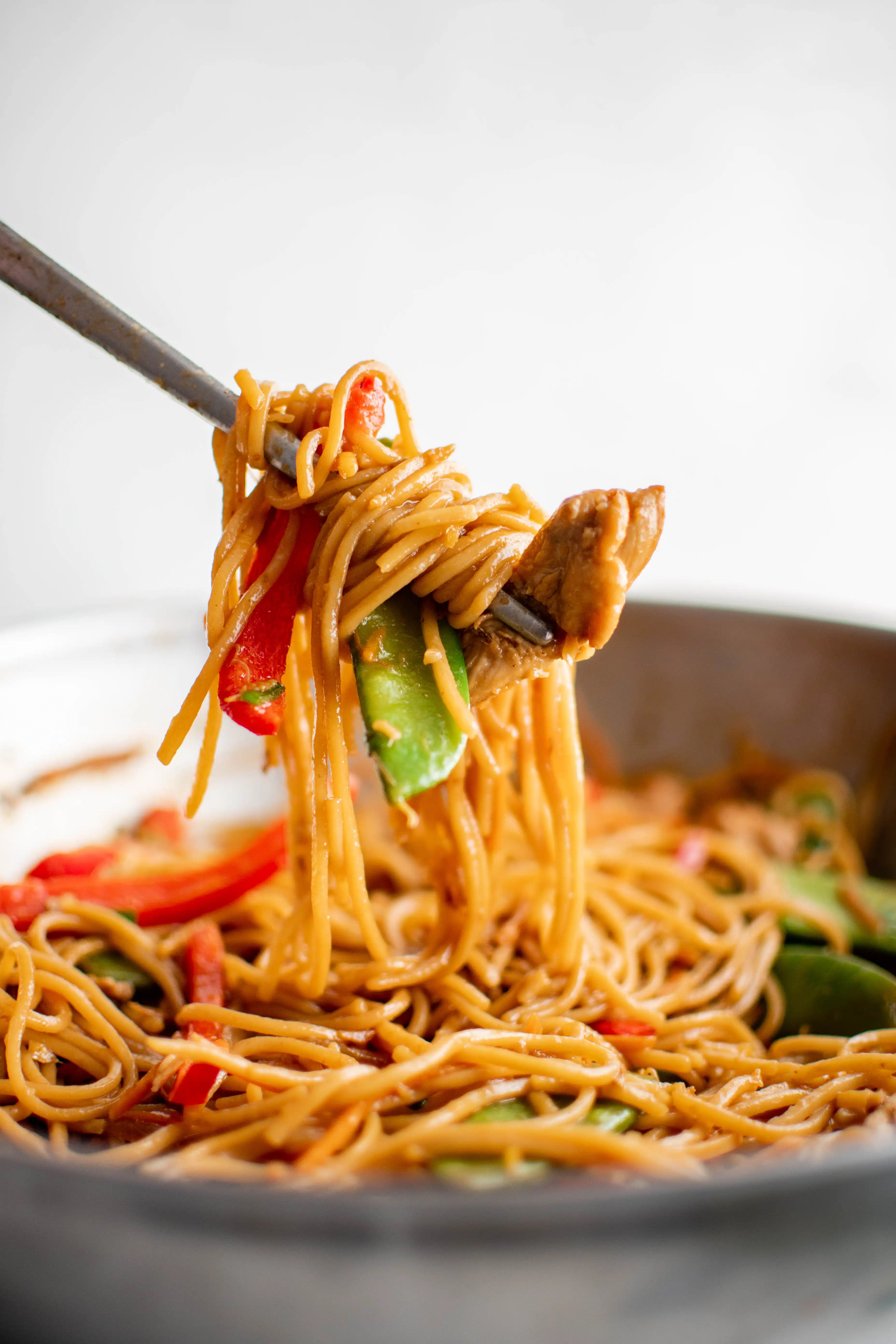 Large metal chopsticks carefully removing a large scoop of chow mein with vegetables and chicken from a steel wok.