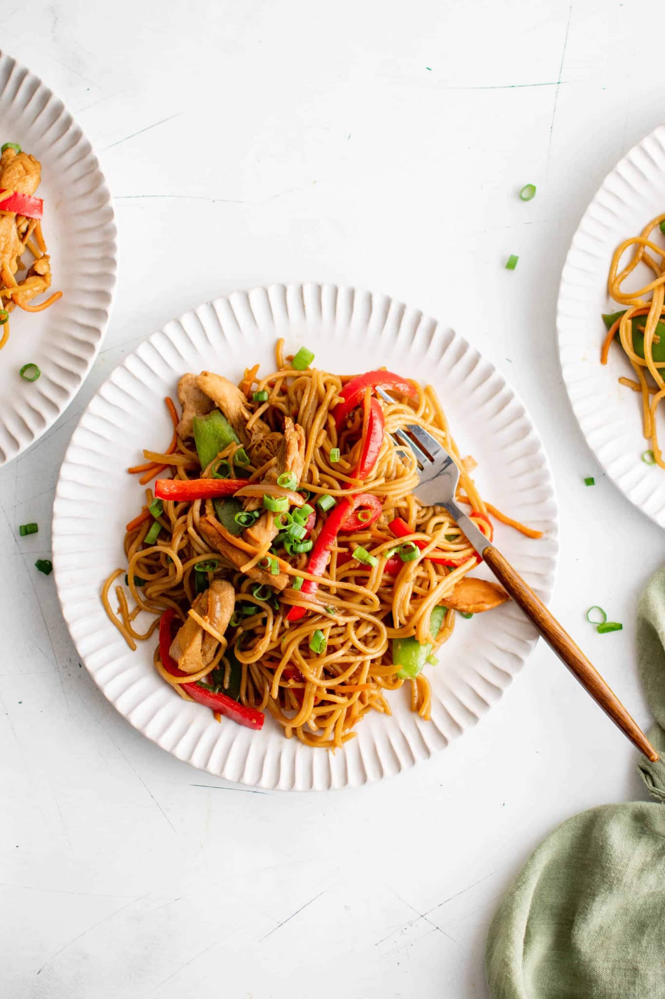 Overhead image of a white plate with crinkled edges filled with a plate of chicken lo mein with onions, carrot, bell pepper, and snow peas and garnished with chopped green onions.