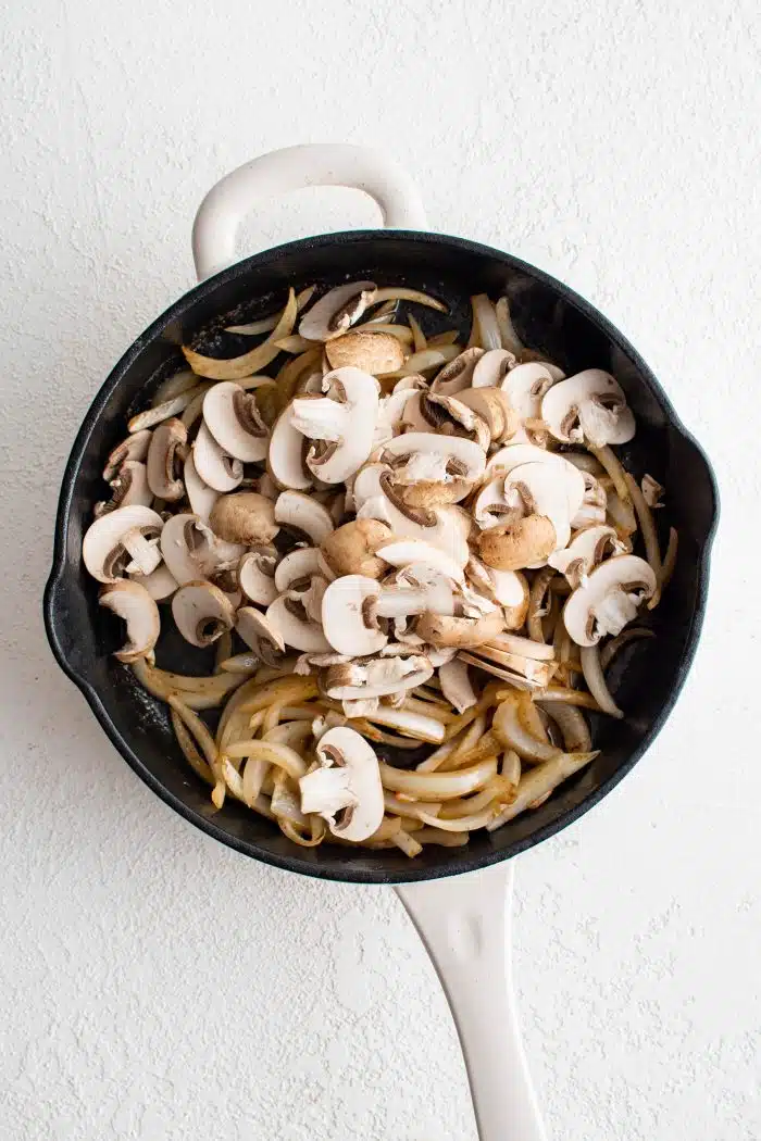 Sliced mushrooms added to a large skillet filled with sauteing onions.