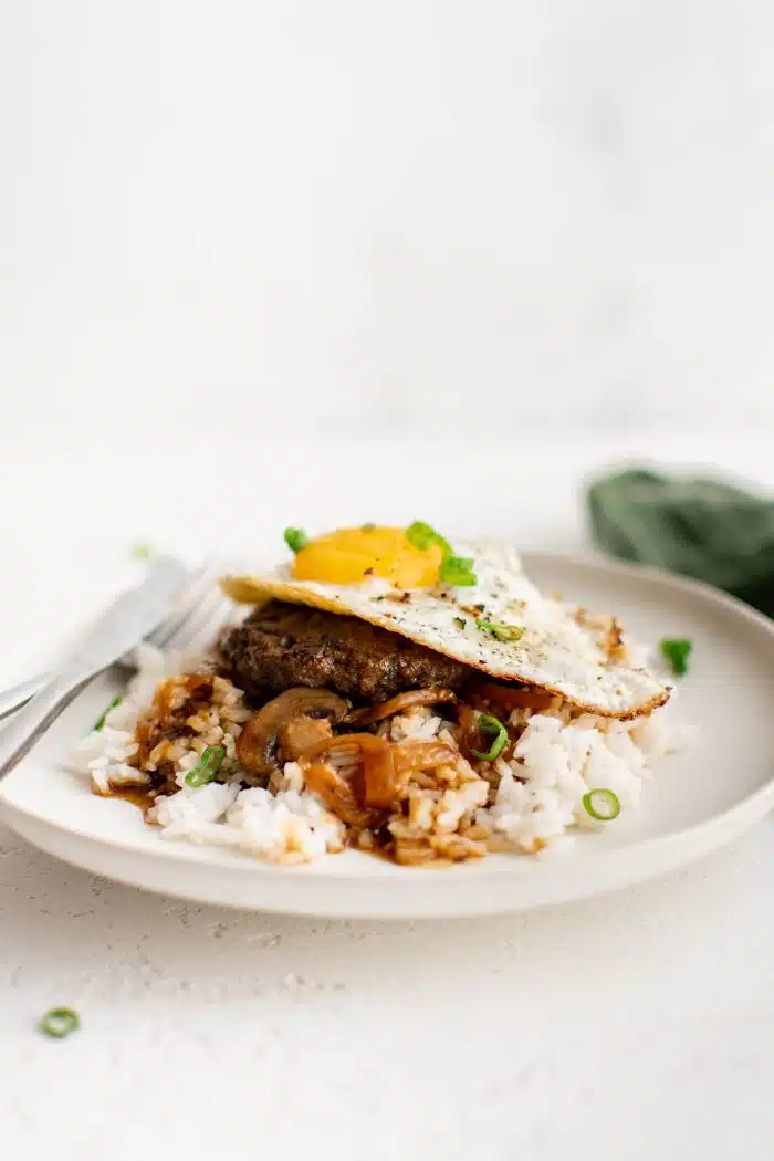 White plate filled with fluffy white rice smothered in an onion and mushroom gravy and topped with a hamburger patty and fried egg and garnished with sliced green onion.