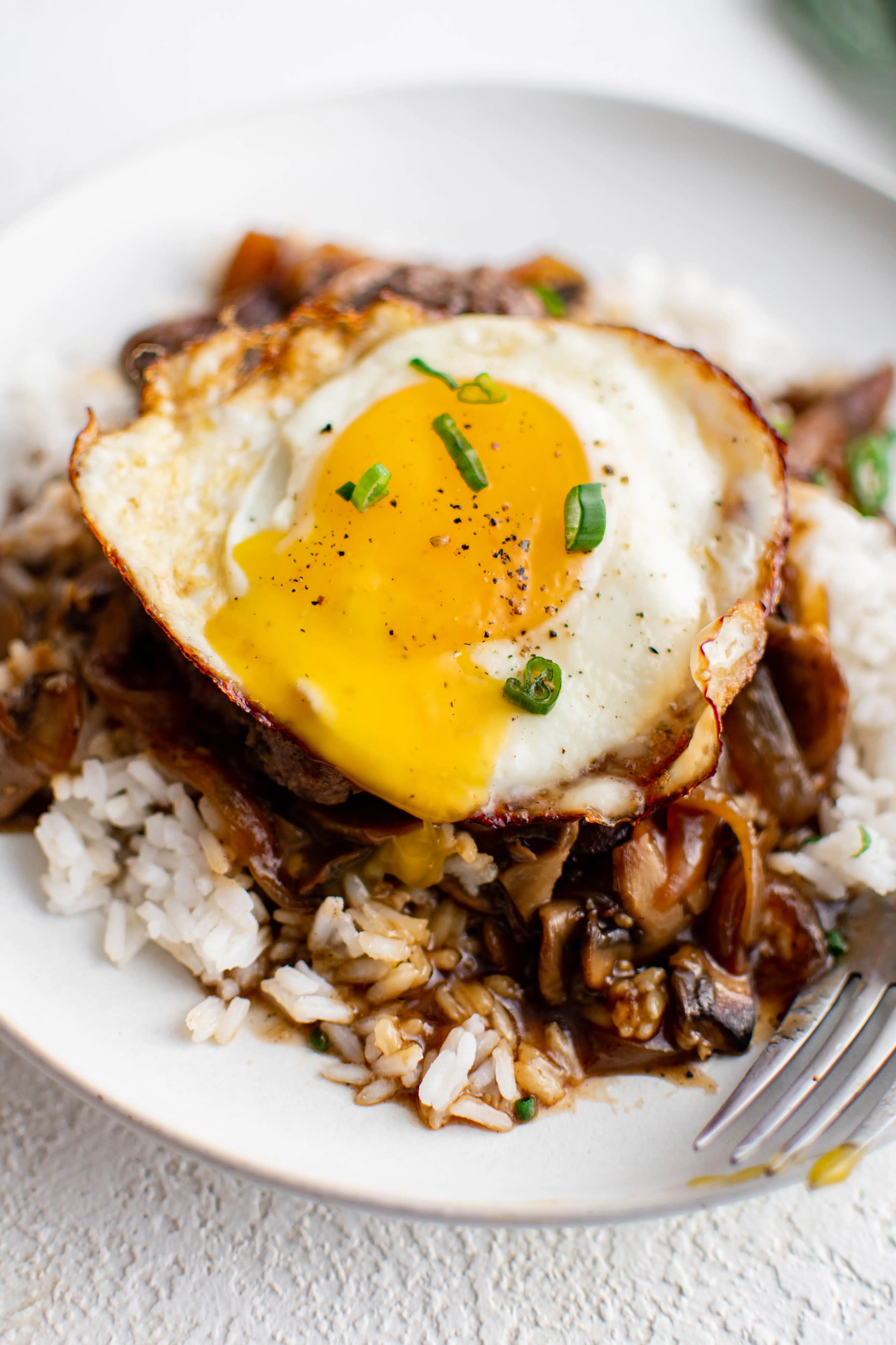 White plate filled with fluffy white rice smothered in an onion and mushroom gravy and topped with a hamburger patty and runny fried egg and garnished with sliced green onion.