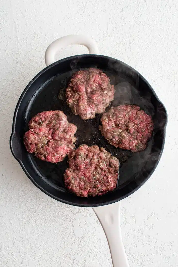 Four hamburger patties cooking in a large skillet.