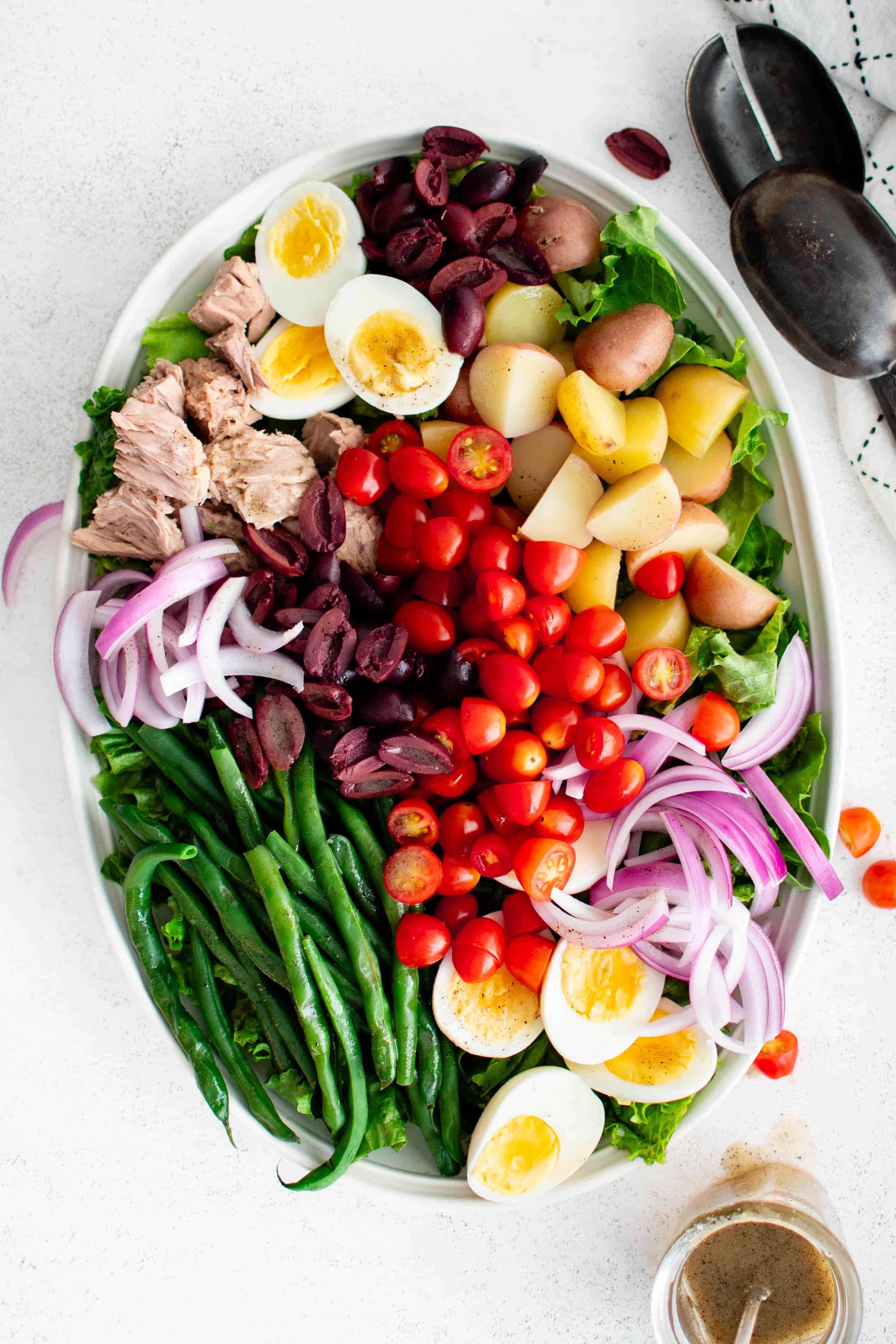 Large oval salad platter artfully displaying a tuna nicoise salad with green beans, hard-boiled eggs, sliced green onions, boiled potatoes, canned tuna, olives, and tomatoes on a bed of lettuce.