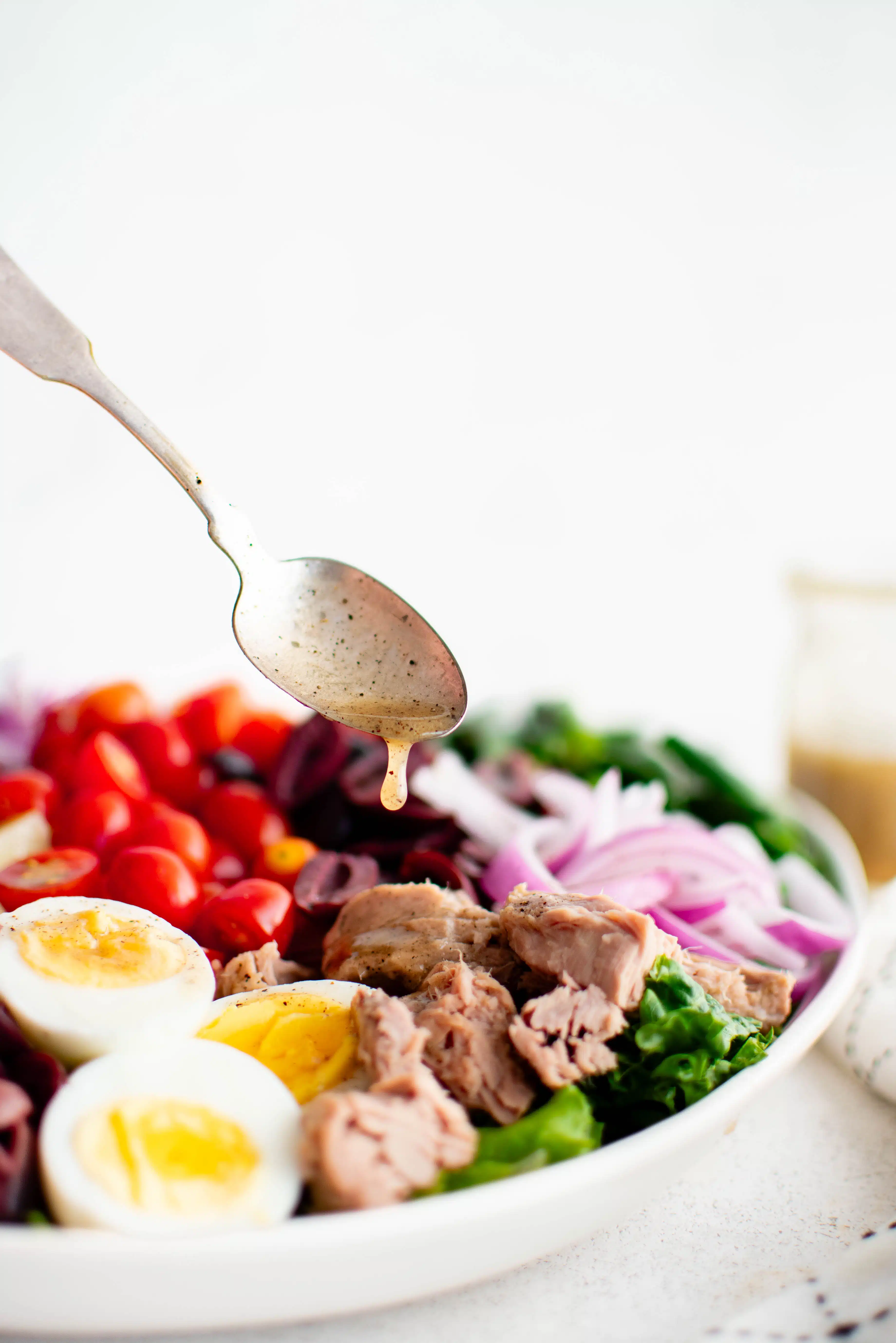 Small spoon drizzling homemade vinaigrette over a large white salad platter artfully presenting nicoise salad.