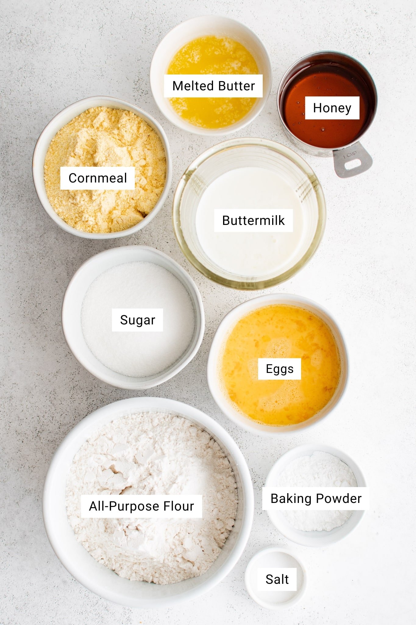 A bright, overhead photo showcasing the measured ingredients for a sweet cornbread recipe. The ingredients are neatly arranged in separate white bowls on a light background. Starting from the top, the image includes a bowl of melted butter and a metal measuring cup filled with honey. Beneath them is a glass bowl of buttermilk, a bowl of beaten eggs, and a bowl of granulated sugar. On the left is a bowl of yellow cornmeal, while at the bottom sits a larger bowl of all-purpose flour, followed by smaller bowls of baking powder and salt. Each ingredient is clearly labeled with text overlays.