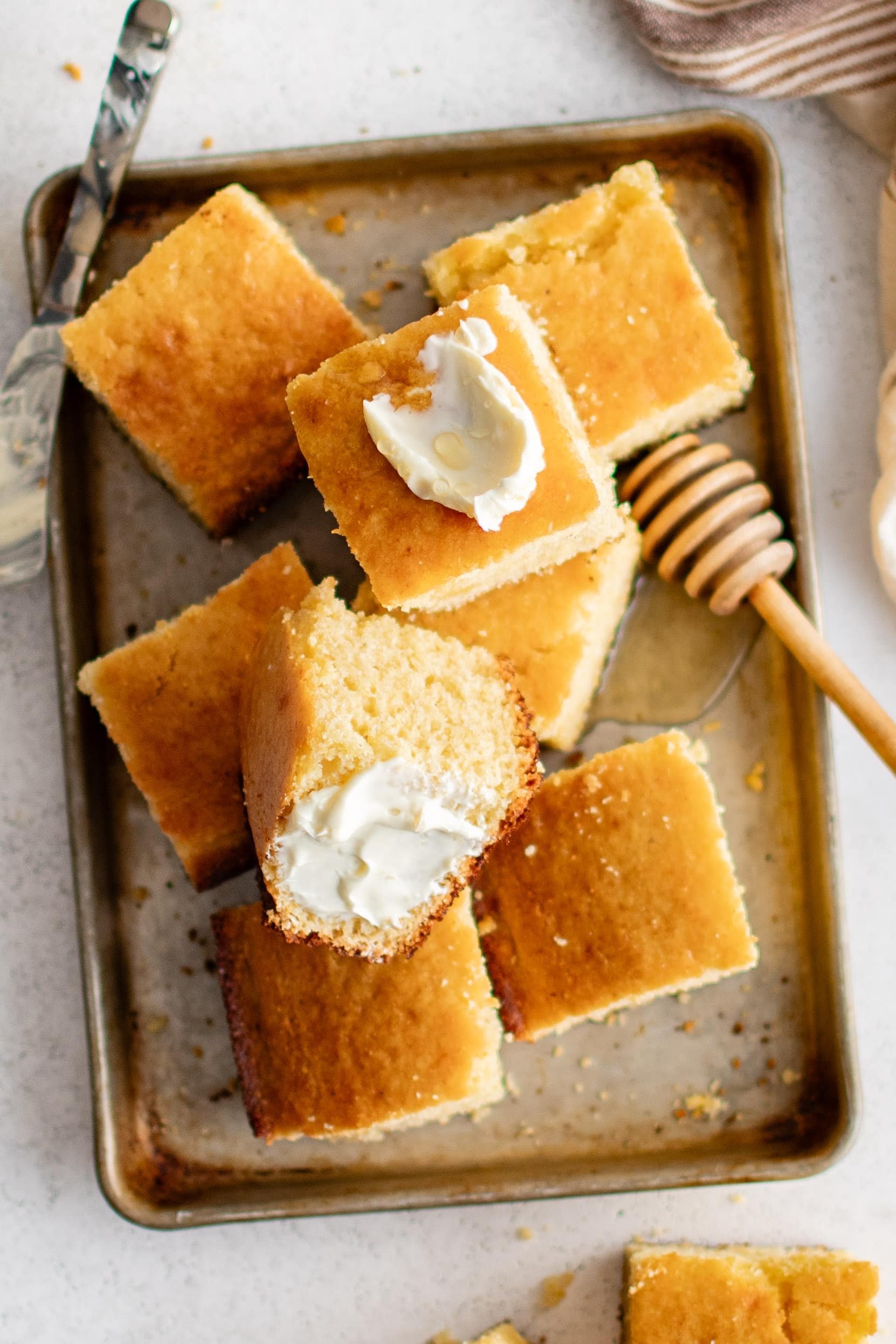 Overhead image of a large rusted baking sheet with a neat pile of cornbread squares, two topped with thick butter and drizzled with butter.