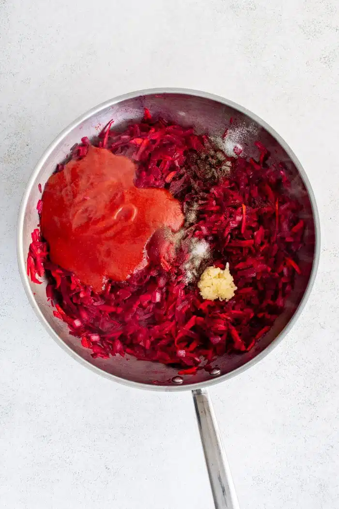 Large stainless steel skillet filled with cooked and softened shredded carrots and beets and diced onion with minced garlic, tomato paste, vinegar, salt, and pepper, and tomato sauce.