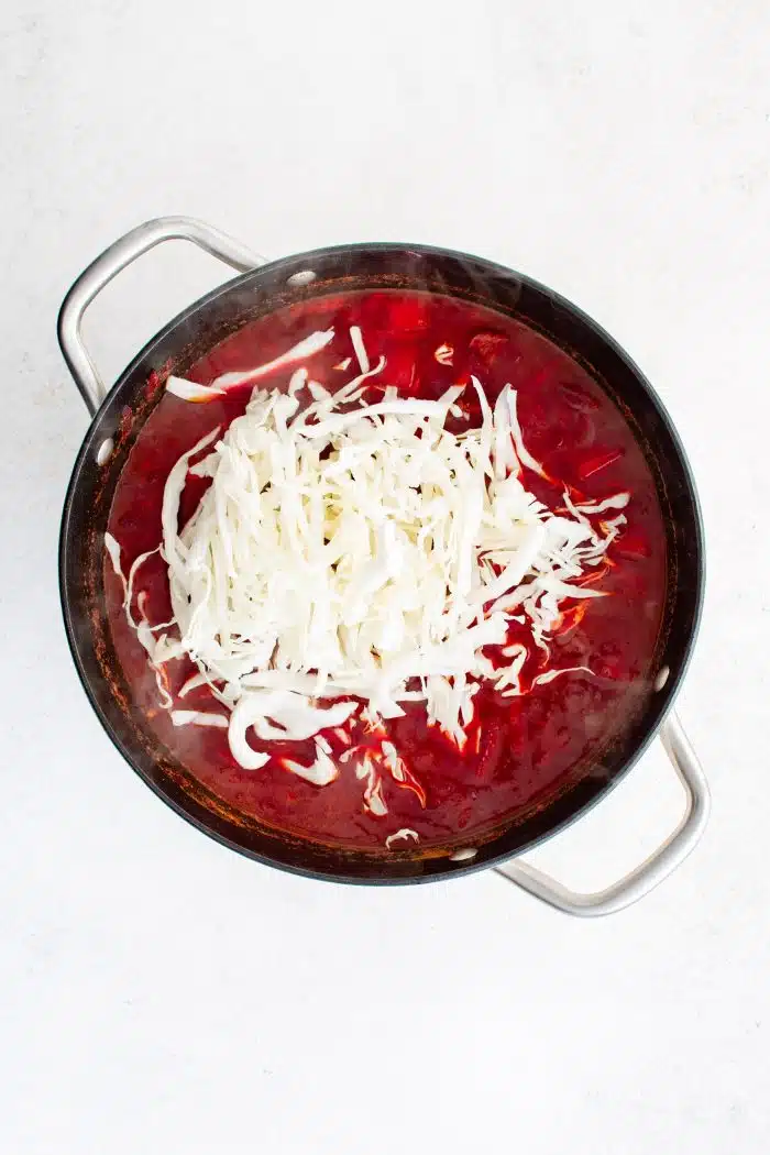 Shredded green cabbage added to a large pot of bright red borscht soup.