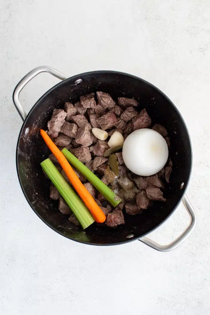 Large pot filled with cooking stew meat, two garlic cloves, one whole onion, two celery sticks, and one carrot.