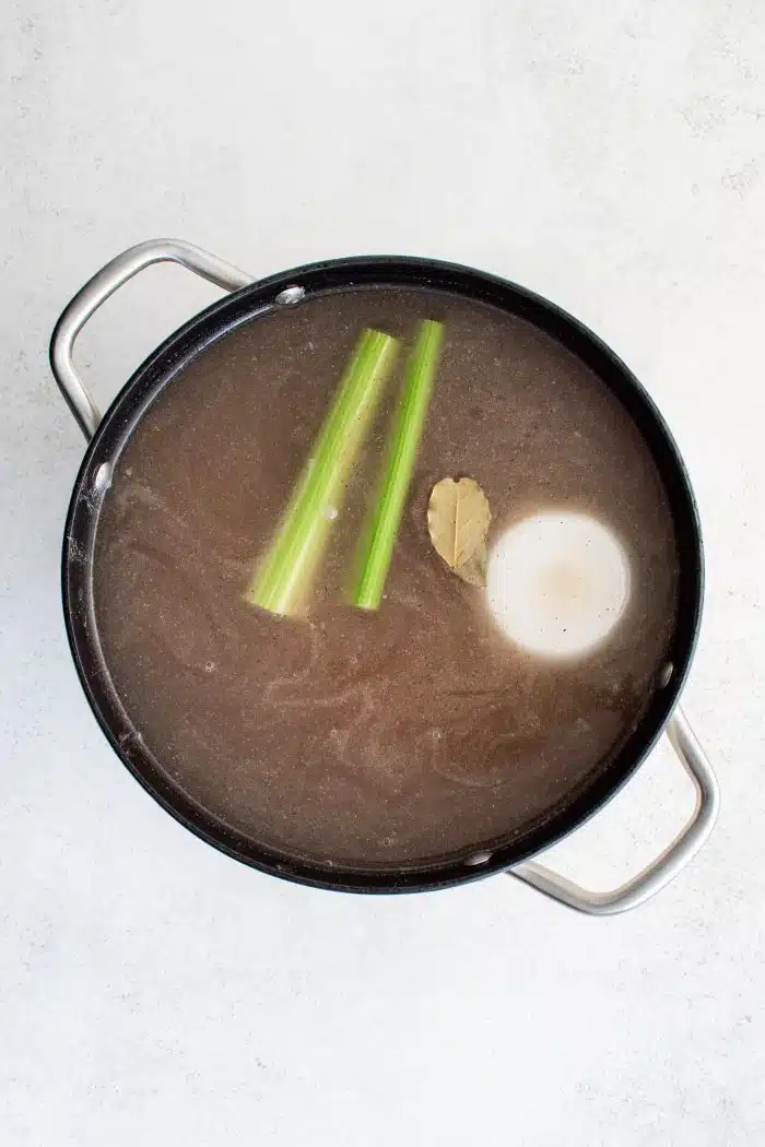 Large soup pot filled with two celery sticks, a bay leaf, and a whole onion floating on top of water that's turned brown from the cooked stew meat sitting on the bottom of the pot.