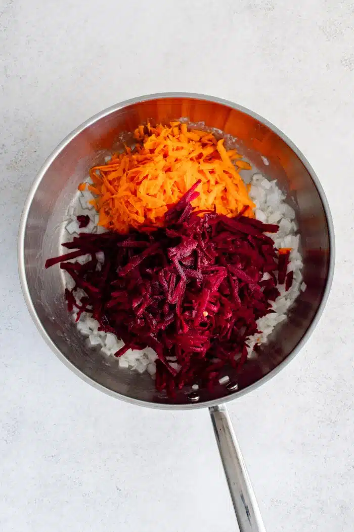 Large stainless steel skillet with softened diced onion covered by shredded raw carrots and shredded raw beets.
