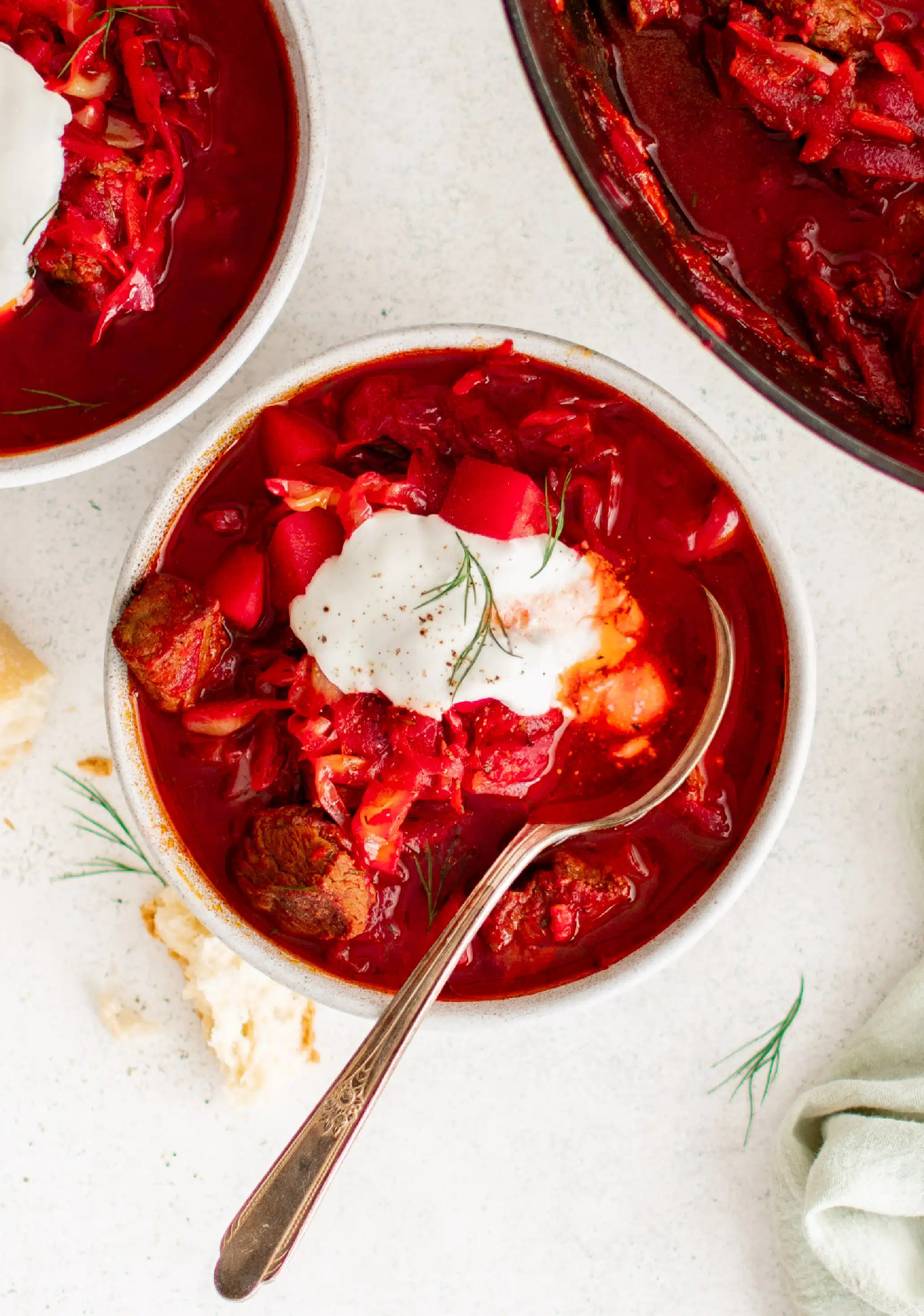 two white bowls filled with borscht soup topped with sour cream set next to a large pot filled with homemade borscht soup.