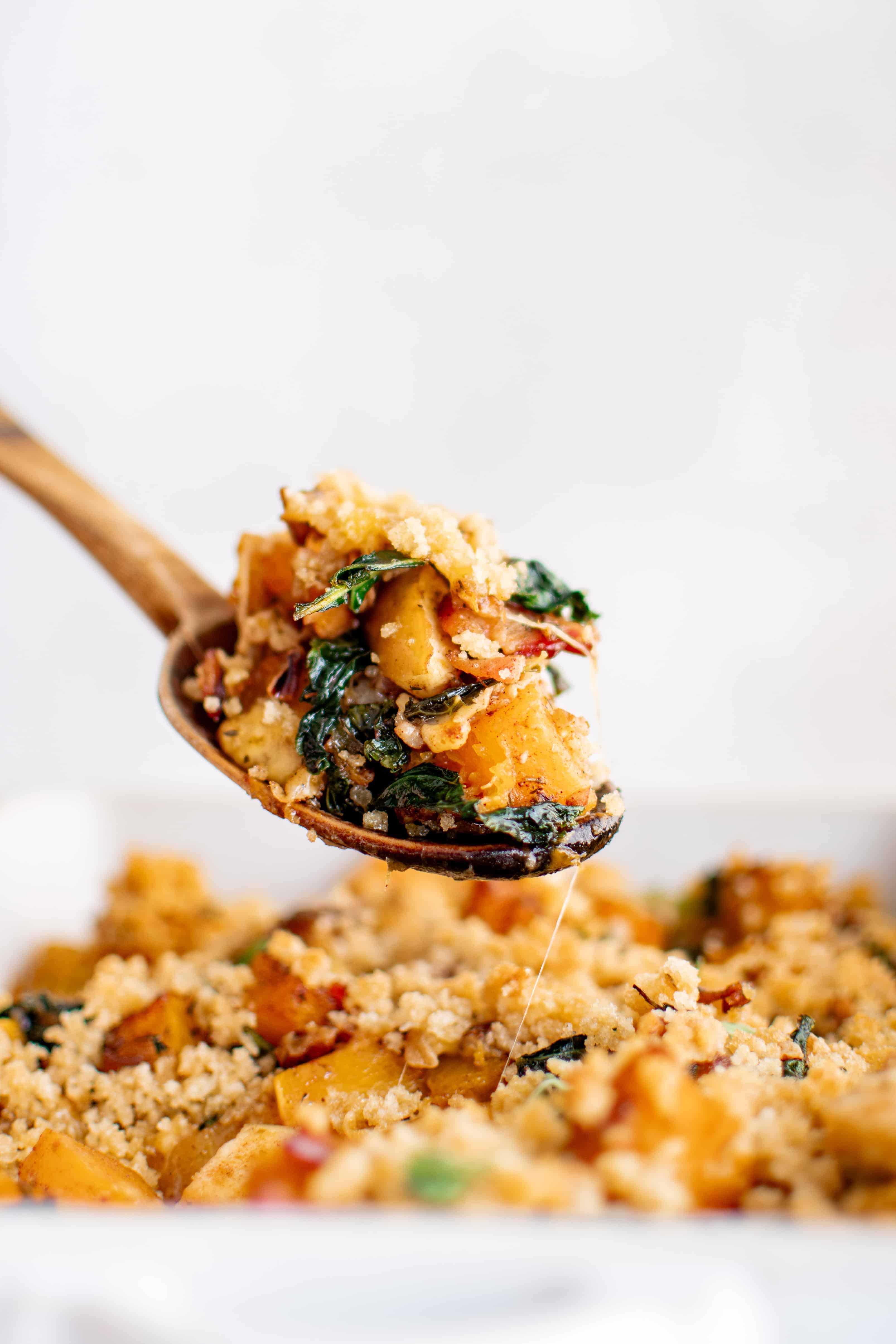 Large serving spoon filled with butternut squash casserole showing everything that it has to offer including kale, pecans, bacon, apple, and cheese.