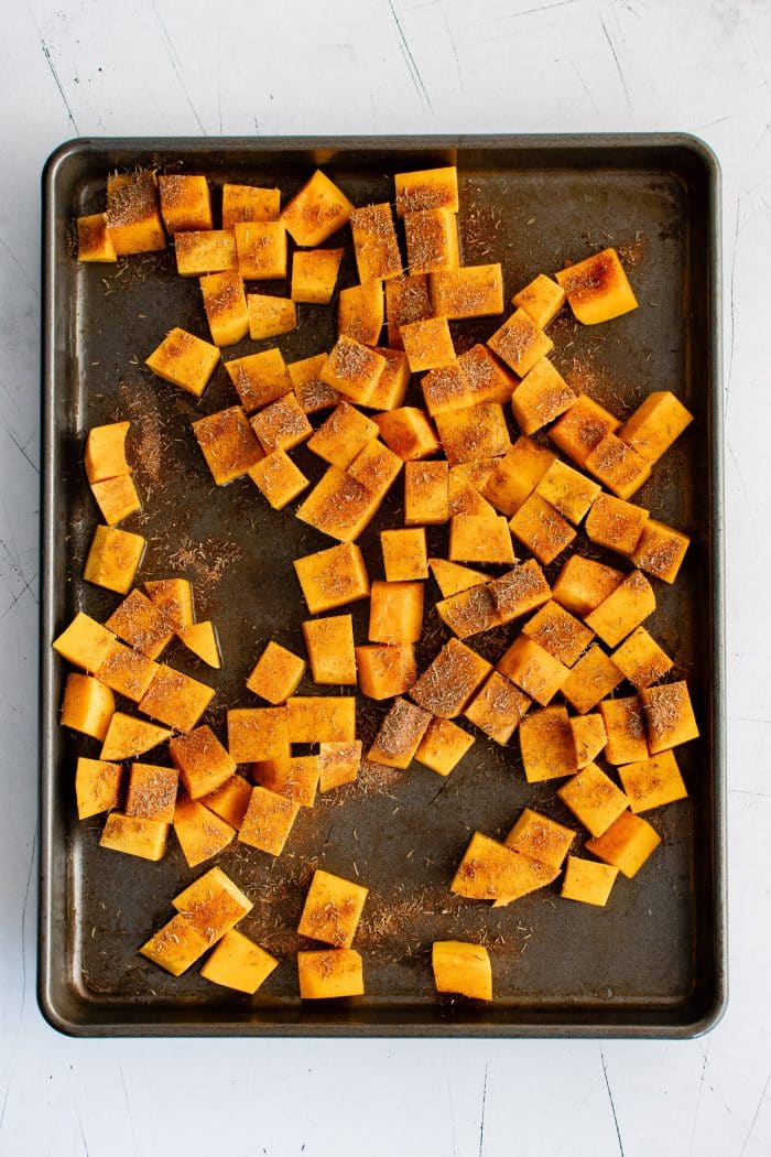 Cubes of uncooked butternut squash on a large baking sheet seasoned with olive oil, dried thyme, salt, dried sage, paprika, ground cinnamon, and nutmeg.