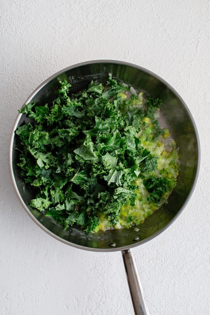 Fresh chopped kale added to a large stainless steel pan filled with sautéed leeks.