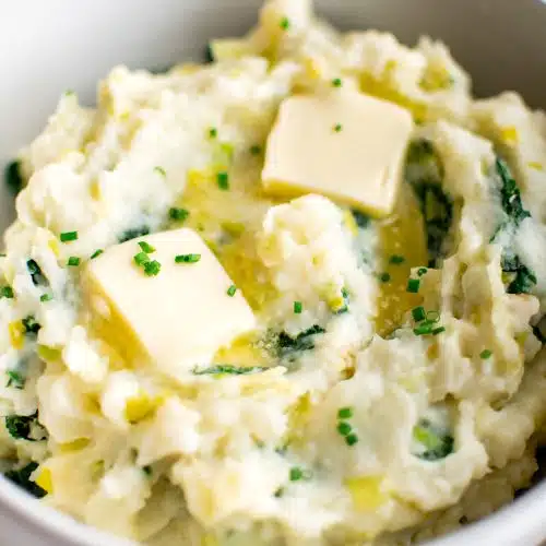 Large white serving bowl filled with colcannon with kale and leeks and topped with to melting pats of butter.