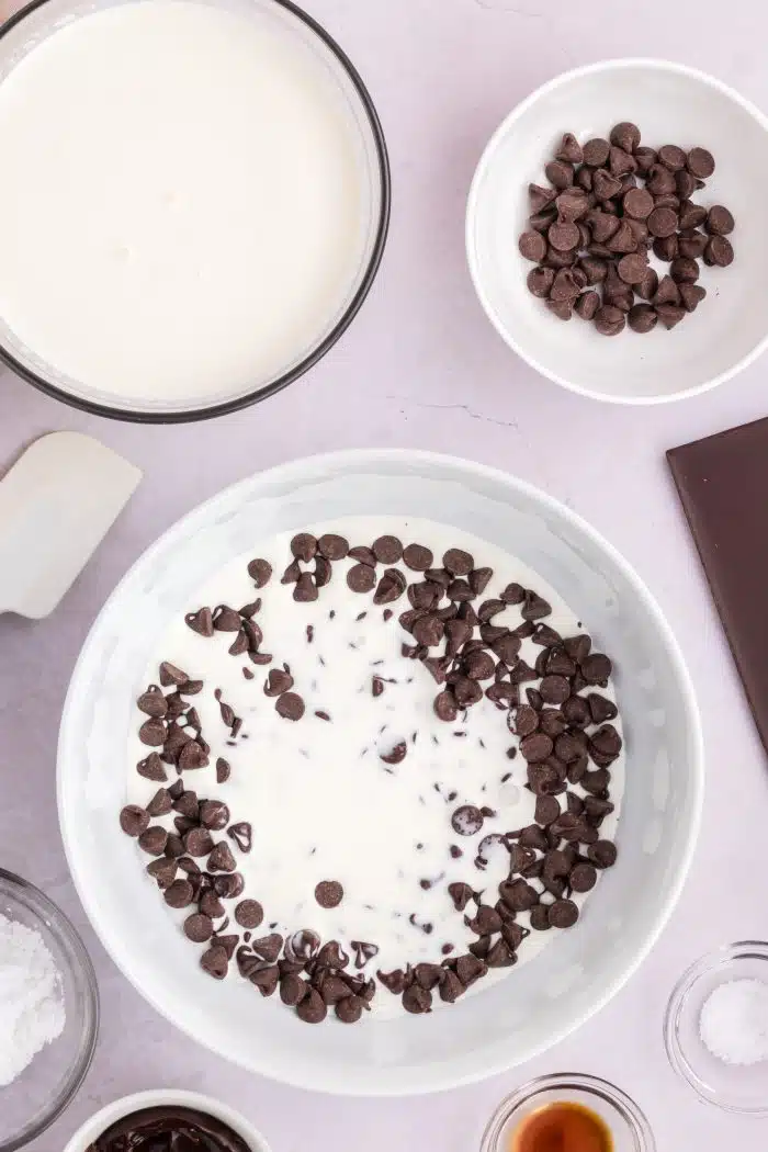 Large white mixing bowl filled with chocolate chips and heavy whipping cream with a bowl of heavy cream and a bowl of chocolate chips set above it.