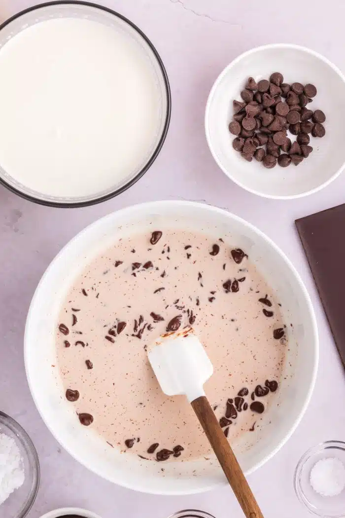 Large white mixing bowl filled with partially melted chocolate chips and heavy whipping cream with a bowl of heavy cream and a bowl of chocolate chips set above it.
