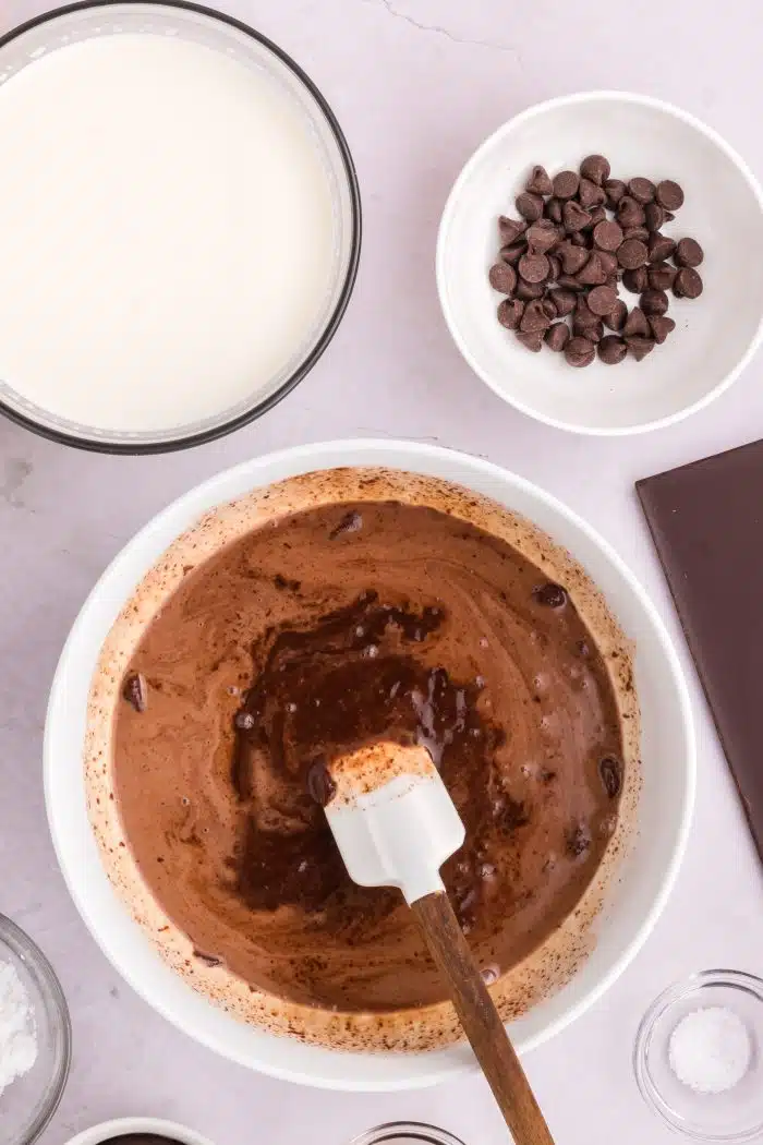 Large white mixing bowl filled with completely melted chocolate chips combined with heavy whipping cream and a bowl of heavy cream and a bowl of chocolate chips set above it.