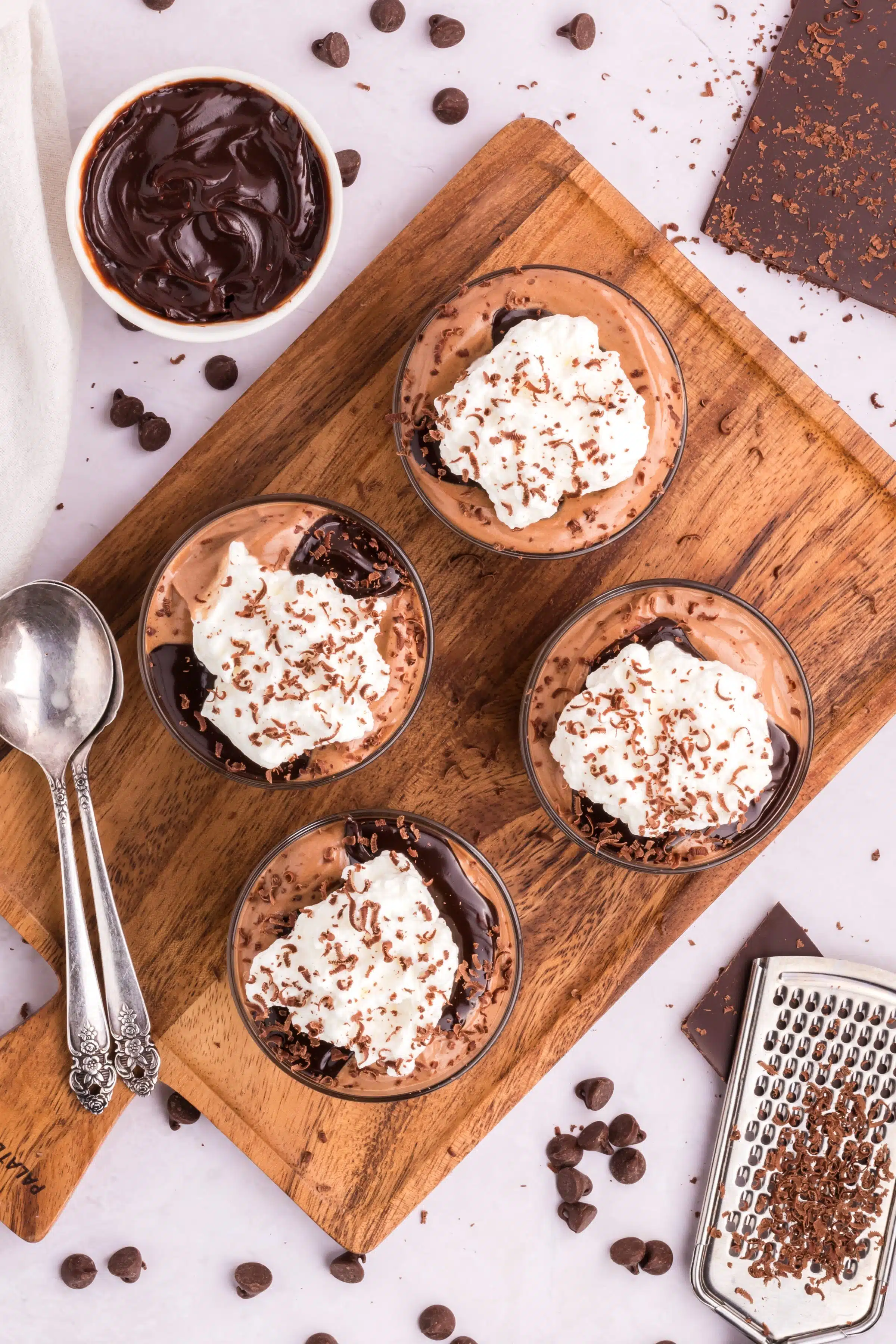 Four glass cups on a rustic cutting board filled with homemade chocolate mousse and topped with chocolate sauce, homemade whipped cream, and shaved chocolate.
