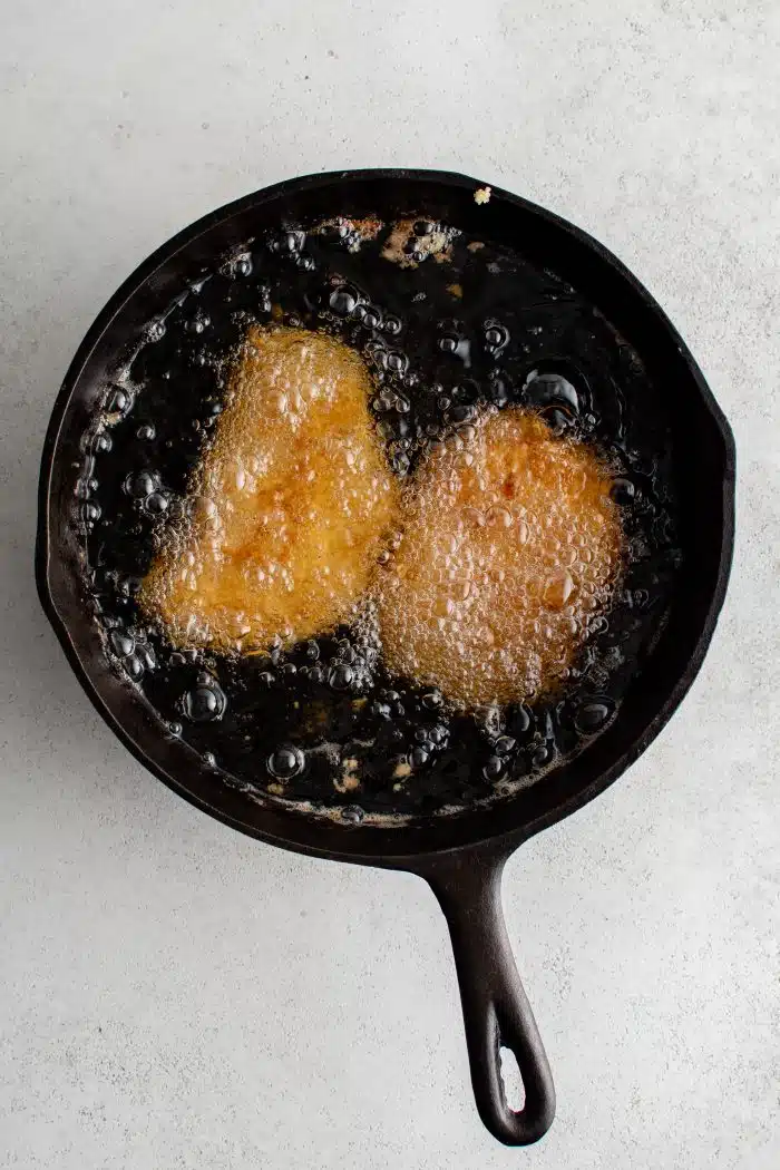 Two breaded pork cutlets deep frying in a large cast-iron skillet.