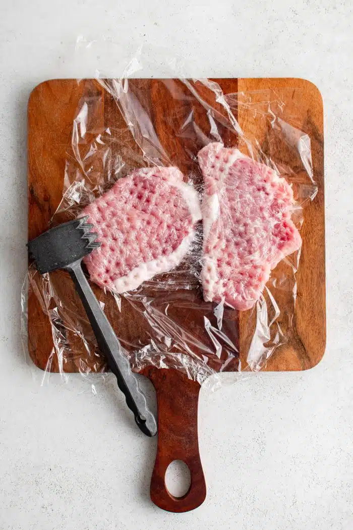 Two pork chops pounded thin with a meat mallet on a wood cutting board under a piece of plastic wrap.