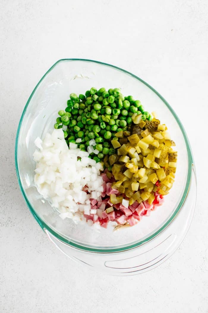 Glass mixing bowl with diced ham, diced dill pickles, diced onion, and green peas.