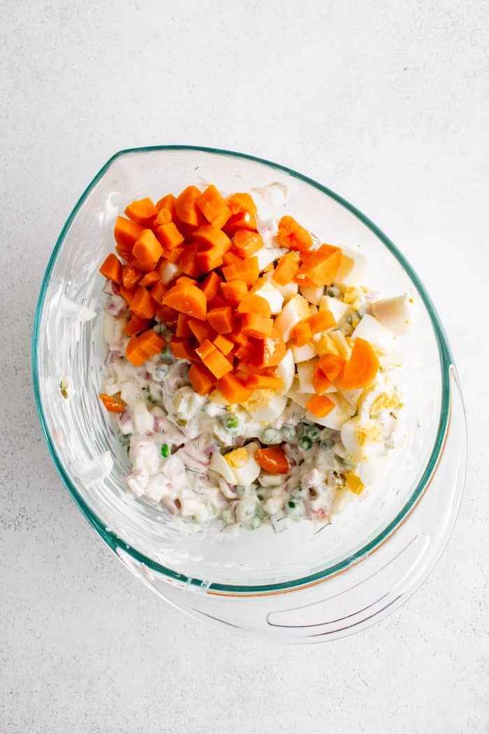 Glass mixing bowl filled with diced ham, diced dill pickles, diced onion, and green peas mixed with dill and mayo dressing and topped with cooked and diced carrots.