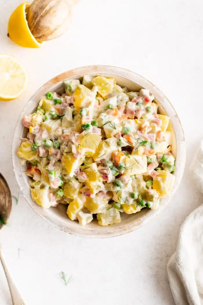 Overhead image of a large serving bowl filled with cold Olivier salad made with cooked potatoes, carrots, hard boiled egg, diced egg, peas, dill pickles, and onion in a creamy mayonnaise dressing.
