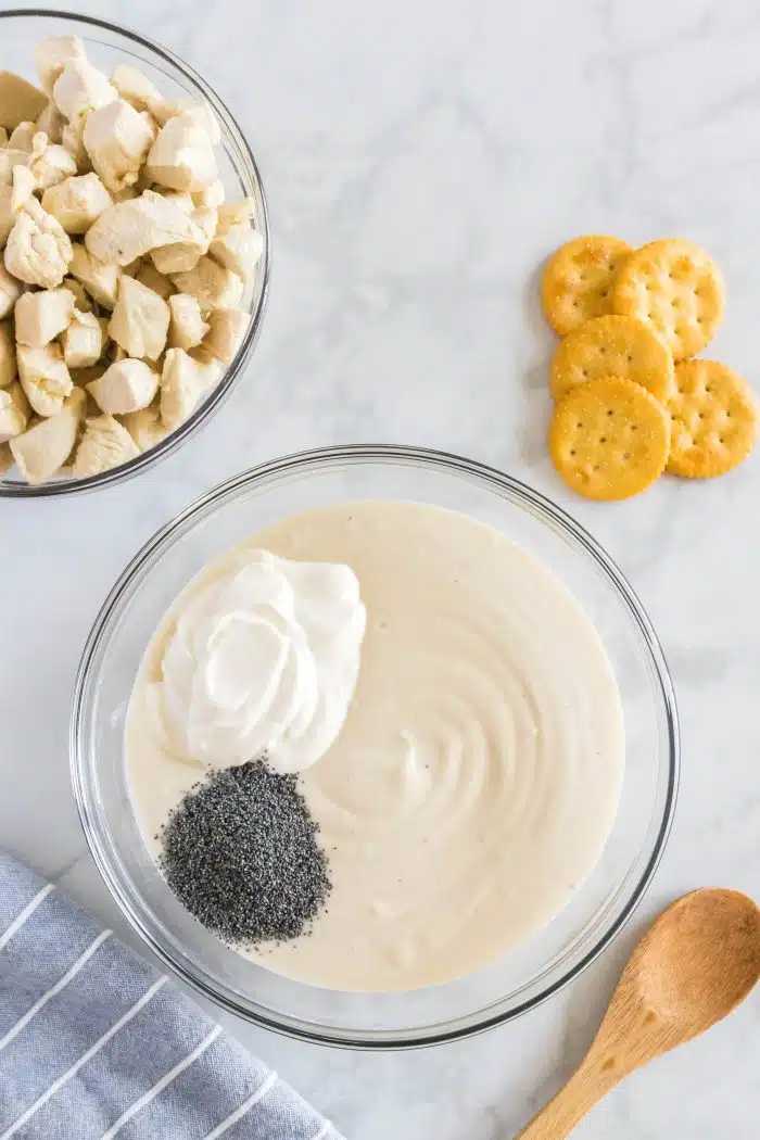 Sour cream and poppy seeds added to a large glass mixing bowl filled with canned cream of chicken soup.