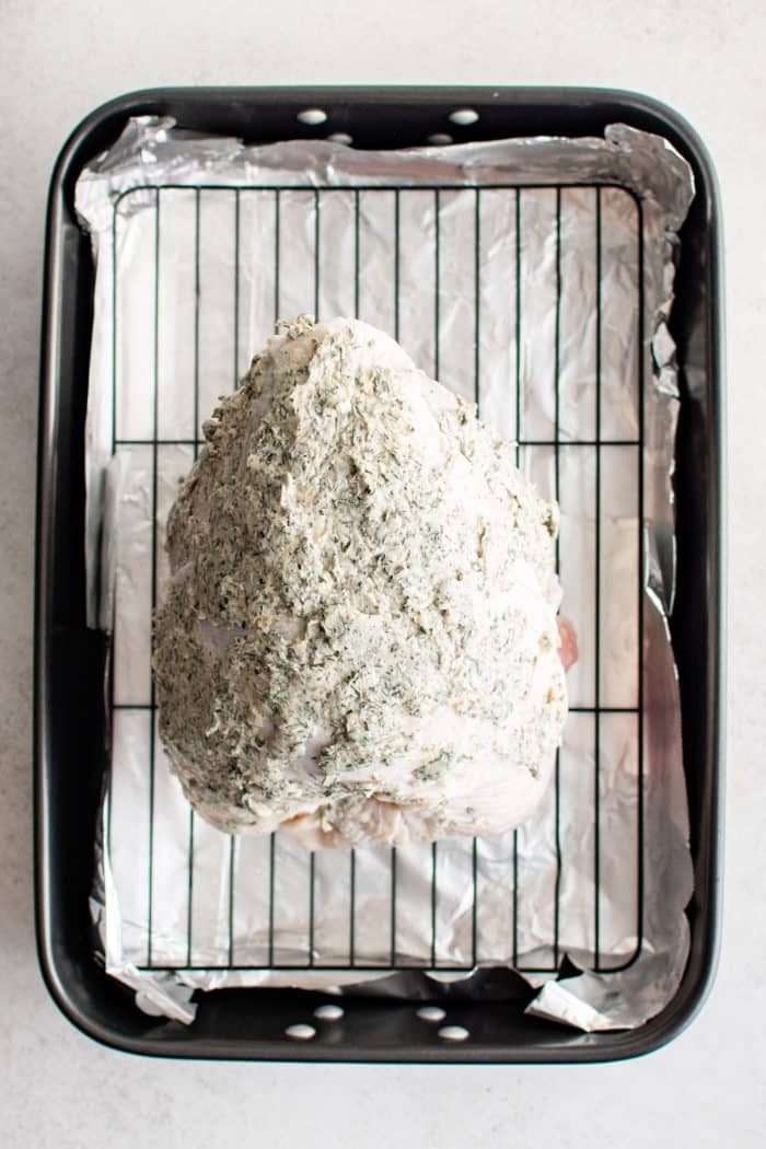 Raw turkey breast coated in herb butter resting on a wire cooking rack set inside a baking sheet lined with aluminum foil.
