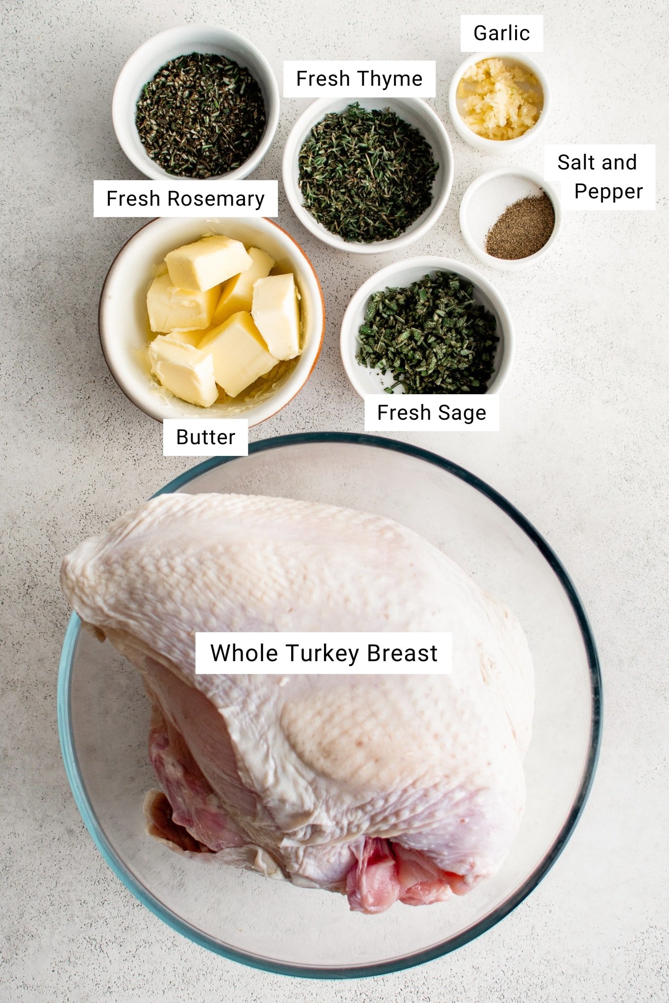 Overhead photo showing a raw bone-in turkey breast with fresh rosemary, thyme, sage, garlic, butter, and seasonings measured out in bowls on a light surface.