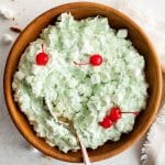 This image features a large wooden bowl filled with fluffy, light green Watergate Salad. The salad is garnished with several bright red maraschino cherries and scattered mini marshmallows. A silver serving spoon is nestled in the salad, ready for scooping. The bowl sits on a white surface with a few stray marshmallows, alongside a textured cream-colored napkin.