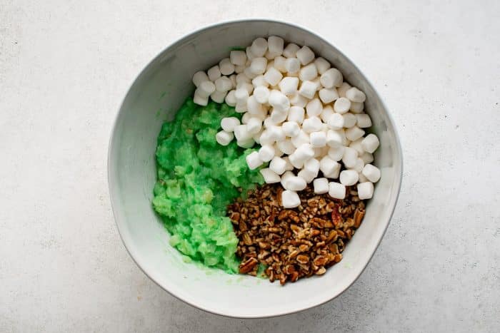 This photo shows a large mixing bowl with the first step of Watergate Salad preparation. One side of the bowl contains a bright green mixture of crushed pineapple and pistachio pudding, while the other sides hold mini marshmallows and chopped pecans. 