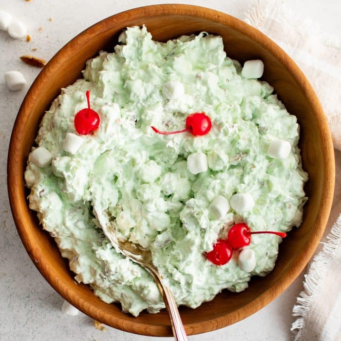 This image features a large wooden bowl filled with fluffy, light green Watergate Salad. The salad is garnished with several bright red maraschino cherries and scattered mini marshmallows. A silver serving spoon is nestled in the salad, ready for scooping. The bowl sits on a white surface with a few stray marshmallows, alongside a textured cream-colored napkin.