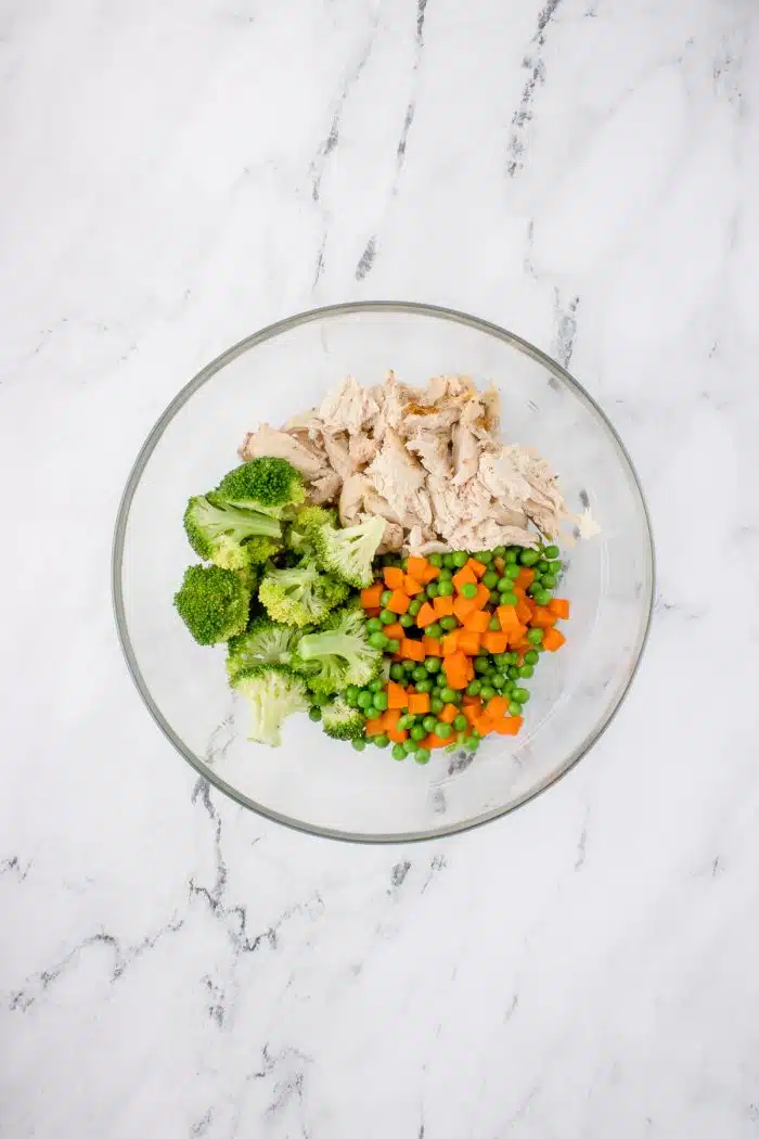 Glass mixing bowl filled with shredded chicken breast, thawed peas and carrots, and steamed broccoli.