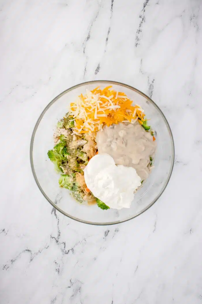 Glass mixing bowl filled with mixed together shredded chicken, thawed peas and carrots, and steamed broccoli with chicken broth, sour cream, spices, shredded cheddar cheese, mozzarella cheese, and cream of mushroom soup.