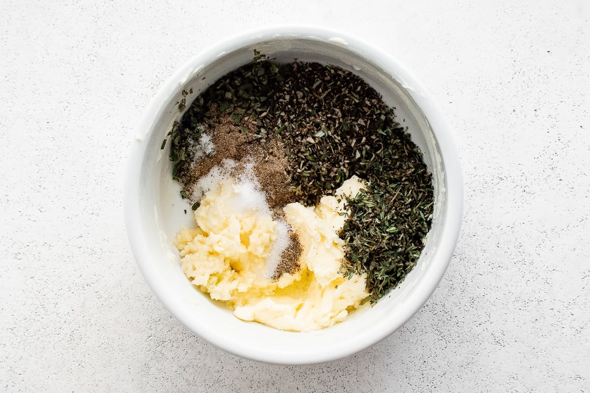 Overhead close-up of butter, chopped herbs, garlic, and seasoning arranged separately in a mixing bowl.