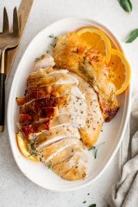 Sliced roast turkey breast with crispy skin arranged on a platter with orange slices and herbs.