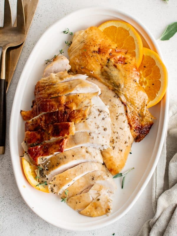 Sliced roast turkey breast with crispy skin arranged on a platter with orange slices and herbs.