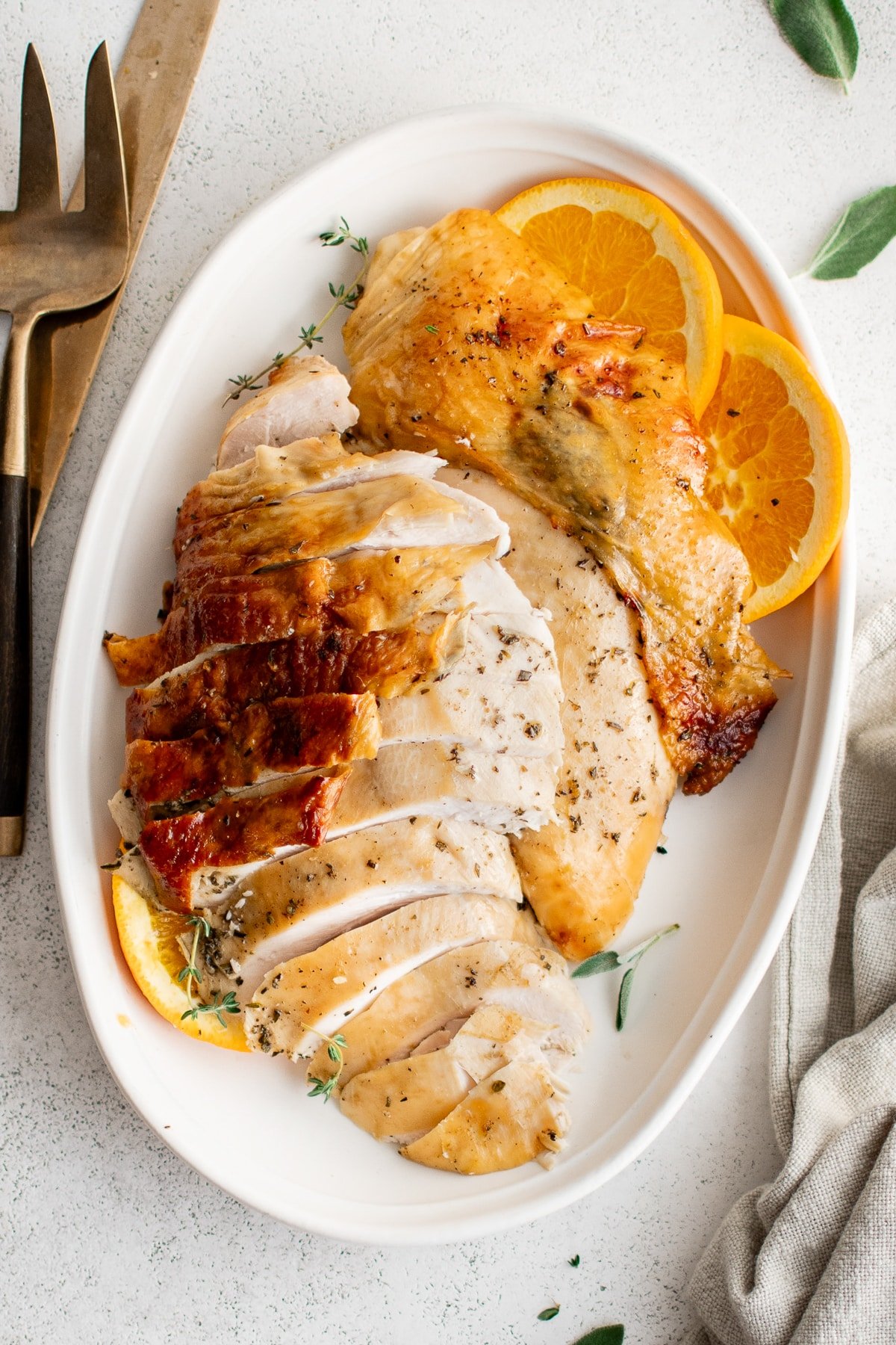 Sliced roast turkey breast with crispy skin arranged on a platter with orange slices and herbs.