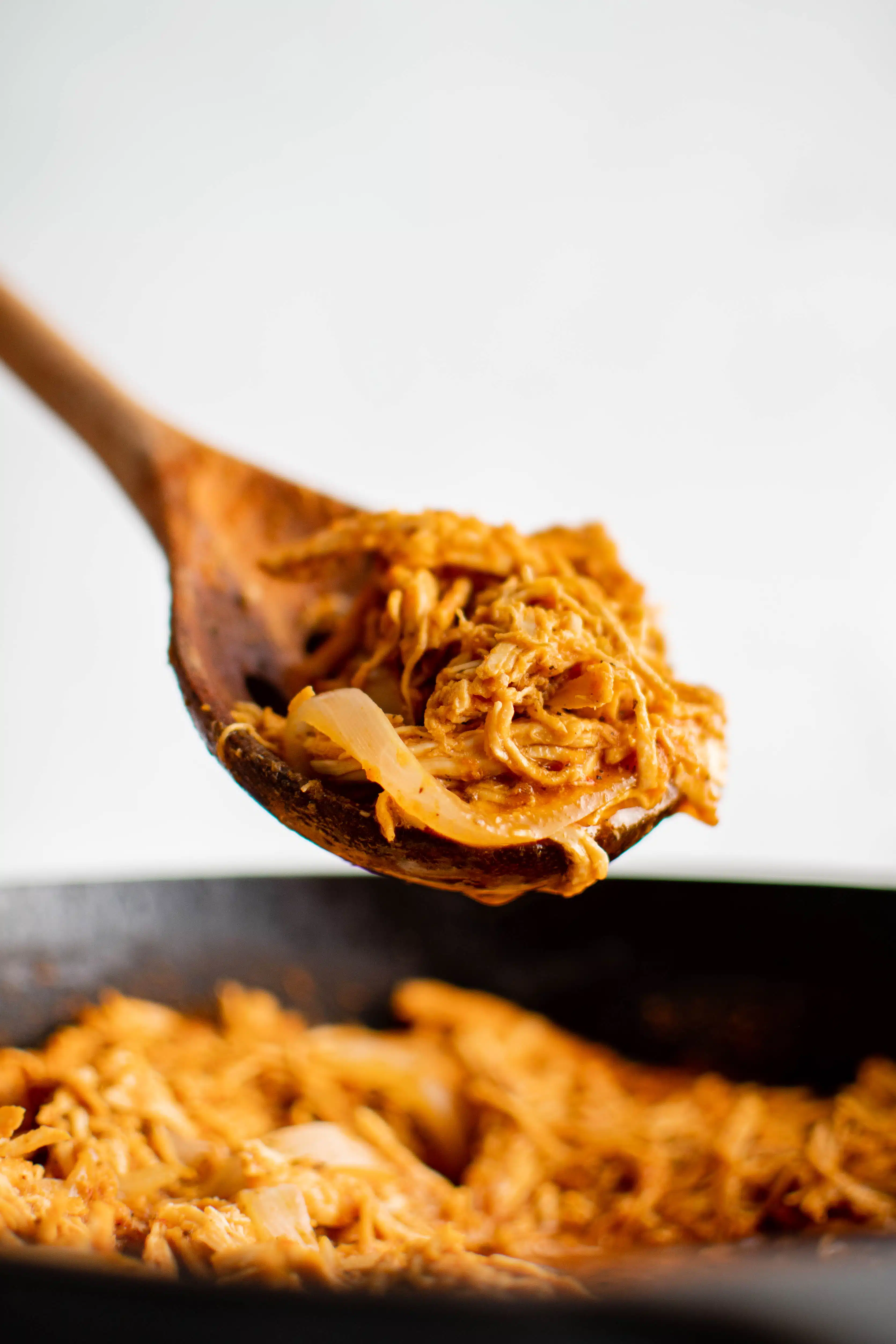 Large wooden serving spoon with a large helping of chicken tinga hovering above a large pan filled with chicken tinga.