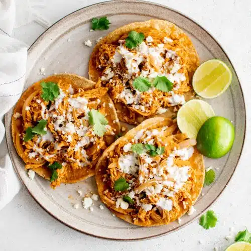 Three chicken tinga tostadas on a round serving plate garnished with crumbled cotija cheese, crema, cilantro, and a side of fresh limes.
