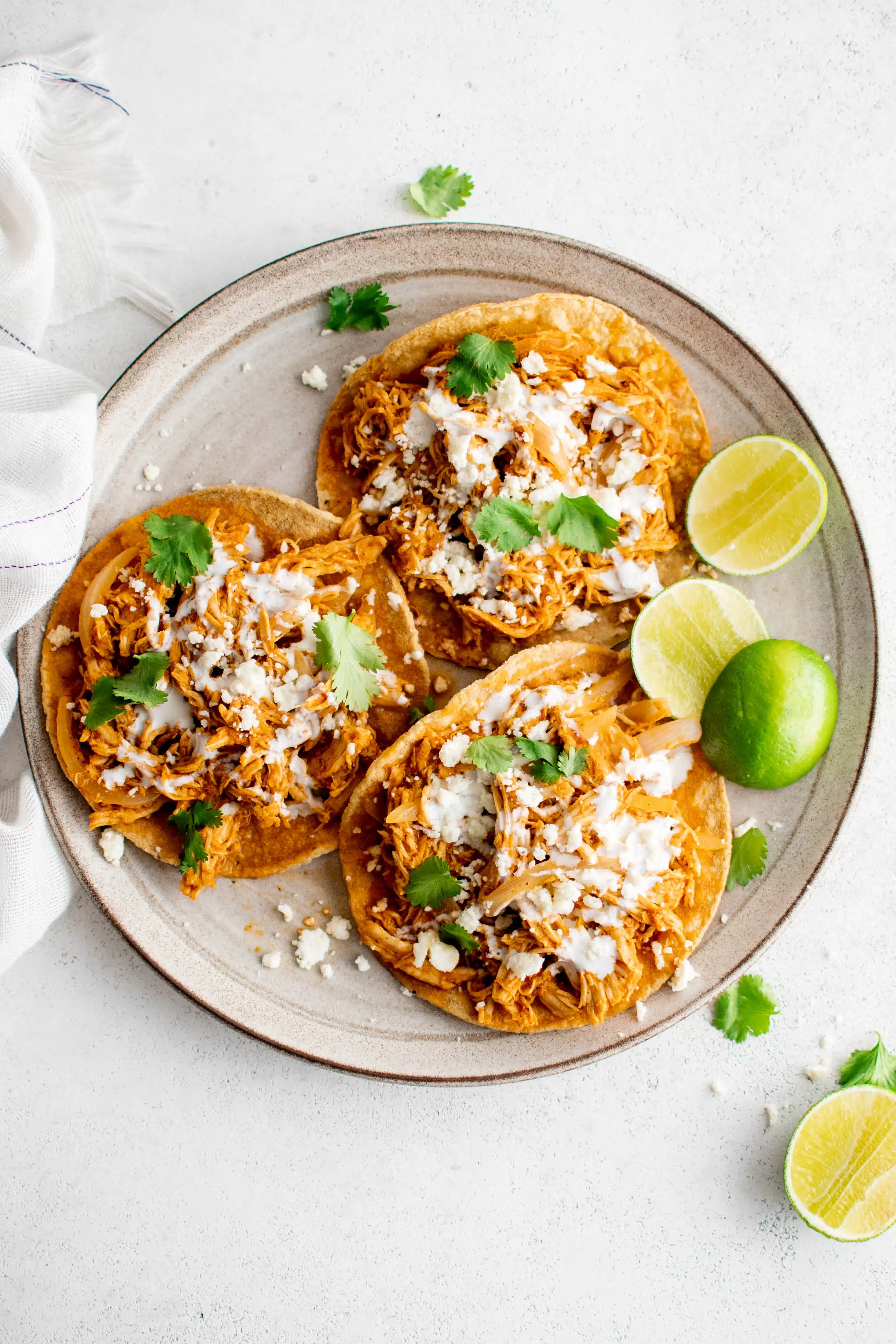 Three chicken tinga tostadas on a round serving plate garnished with crumbled cotija cheese, crema, cilantro, and a side of fresh limes.