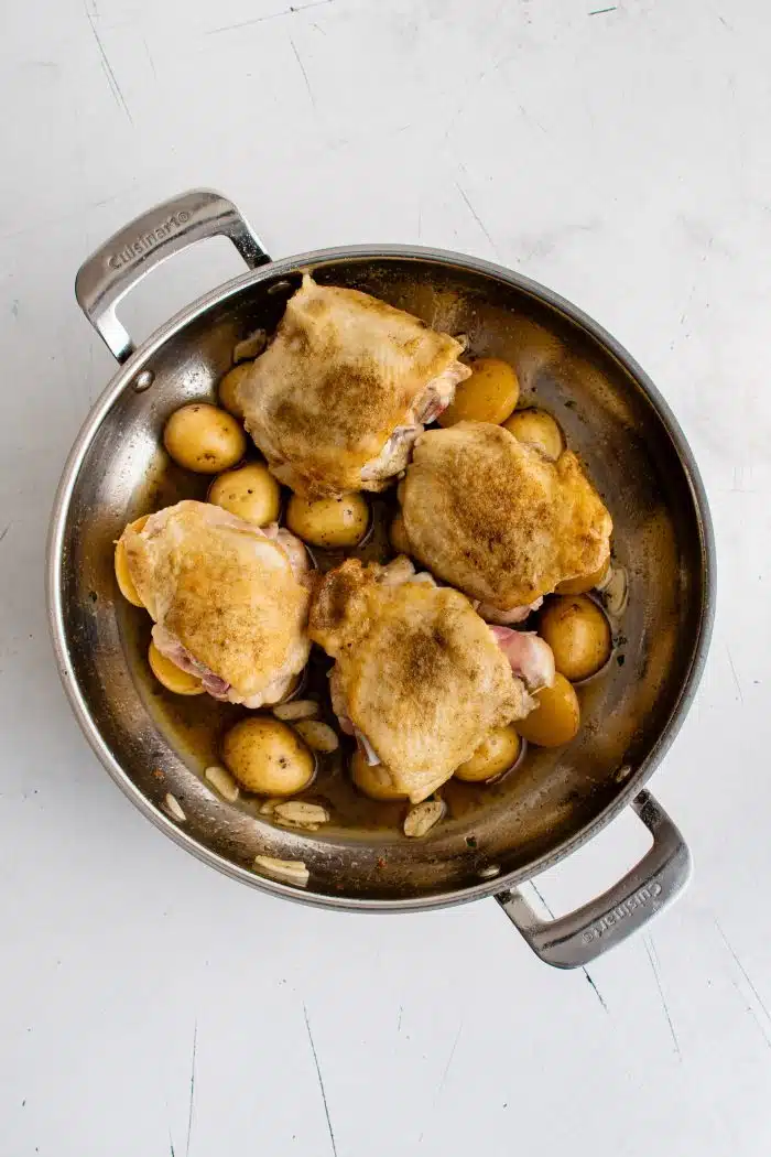 Seared chicken thighs placed on top of halved baby potatoes in a lemon garlic white wine sauce cooking in a large pan.