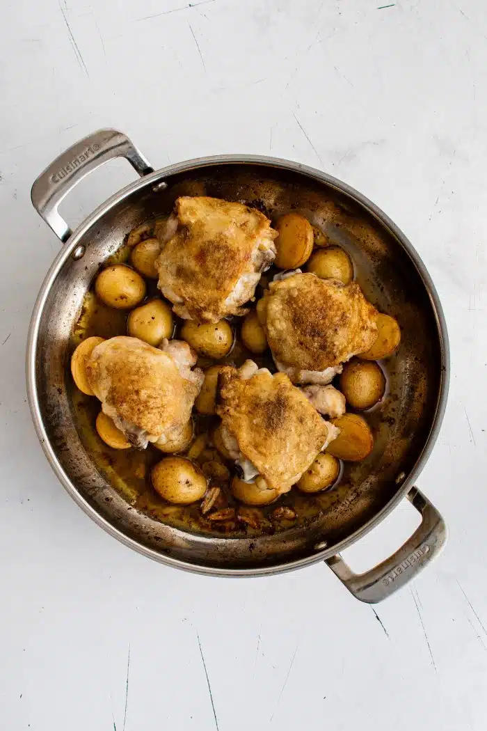 Roasted chicken thighs on top of halved baby potatoes in a lemon garlic white wine sauce cooking in a large pan.