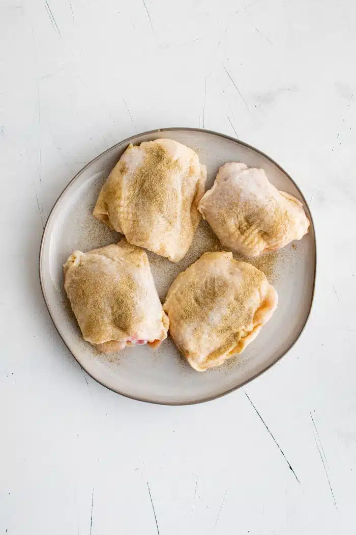 Four bone-in skin-on chicken thighs on a white plate seasoned with salt and poultry seasoning.