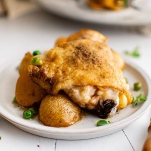 Small white dinner plate filled with a seared and oven-roasted chicken thigh, halved potatoes, and green peas drizzled with a fresh lemon garlic and white wine sauce.