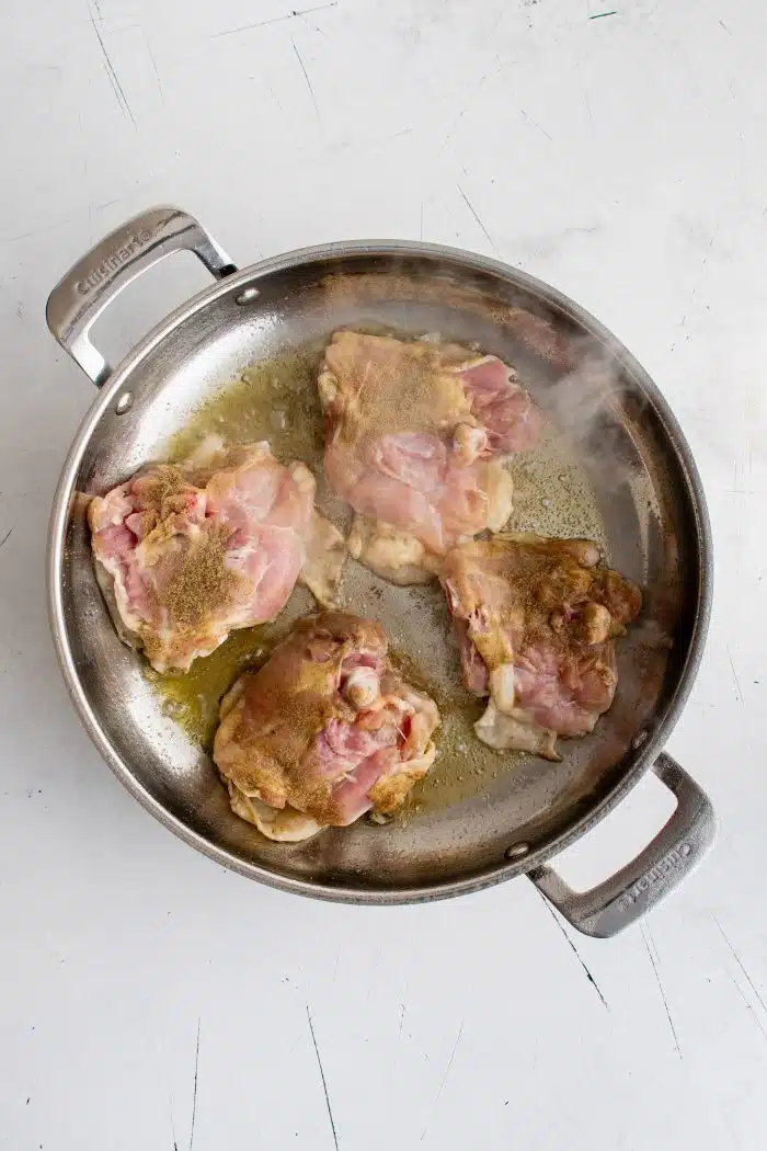 Four large bone-in skin-on chicken thighs being seared in a large skillet skin-side-down.