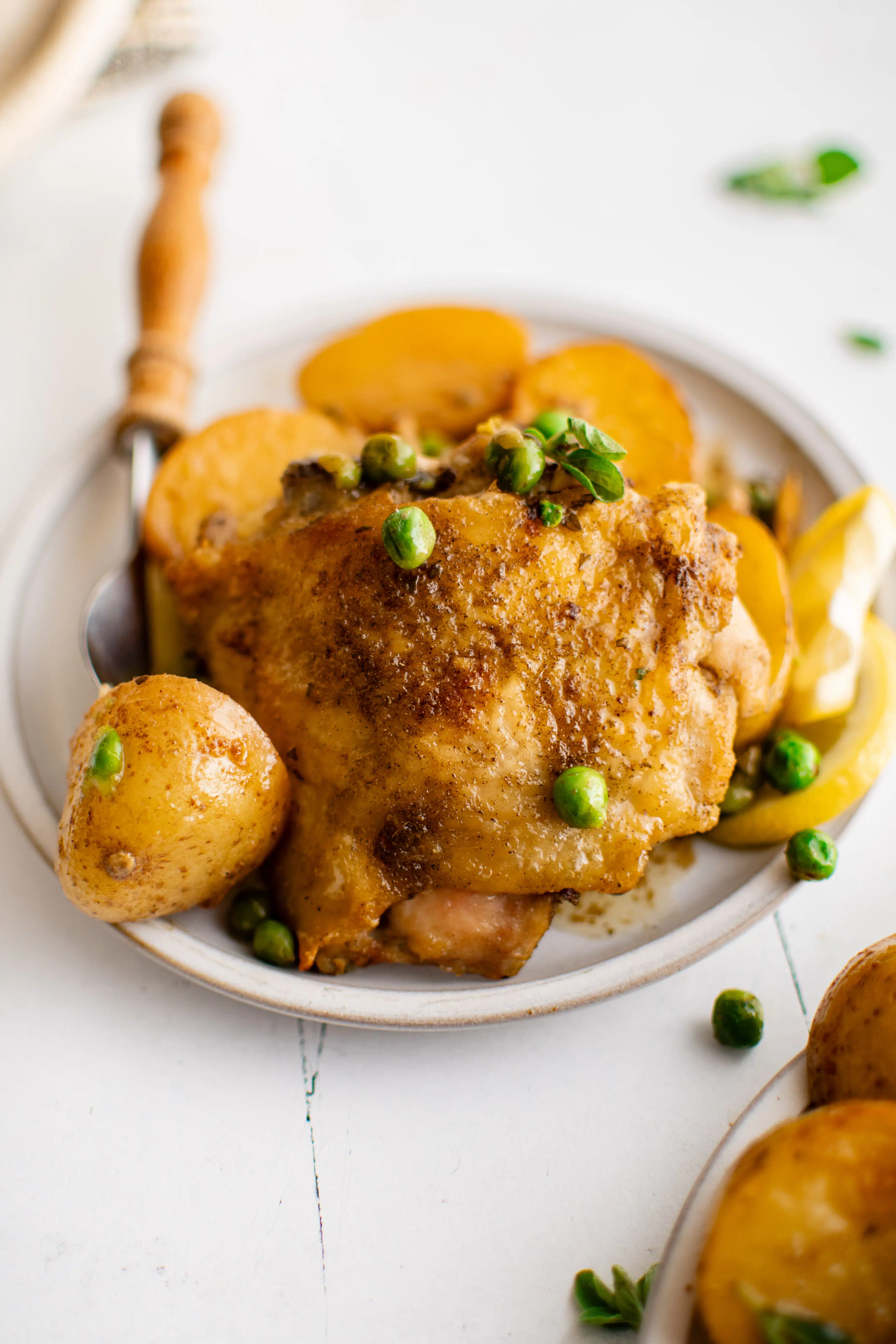 Small white dinner plate filled with a seared and oven-roasted chicken thigh, halved potatoes, and green peas drizzled with a fresh lemon garlic and white wine sauce.